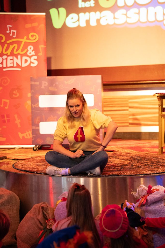 Presentatrice zit op het podium en praat met kinderen.